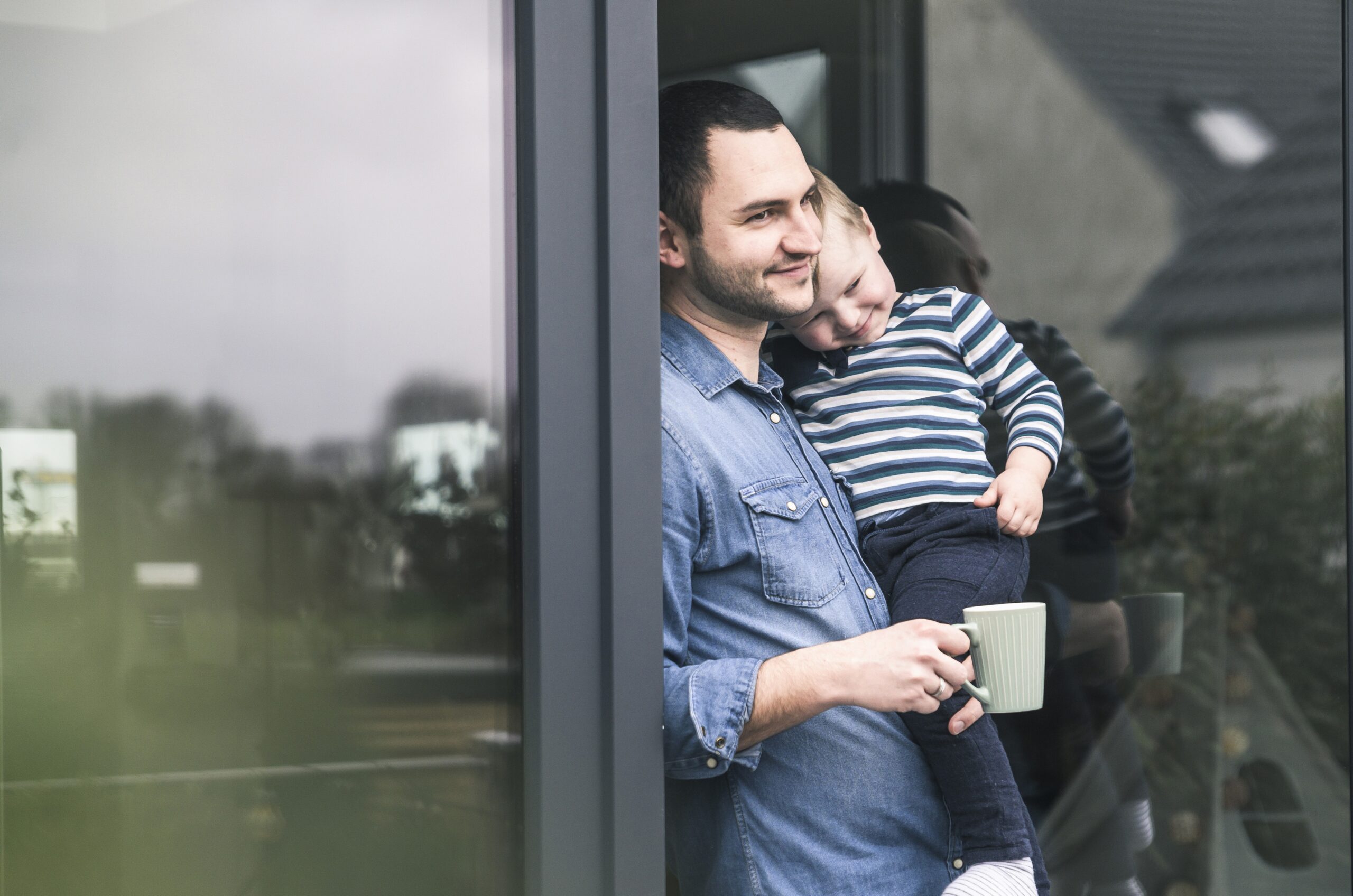 Father with cup of coffee carrying son at terrace door at home after finalising home mover mortgage